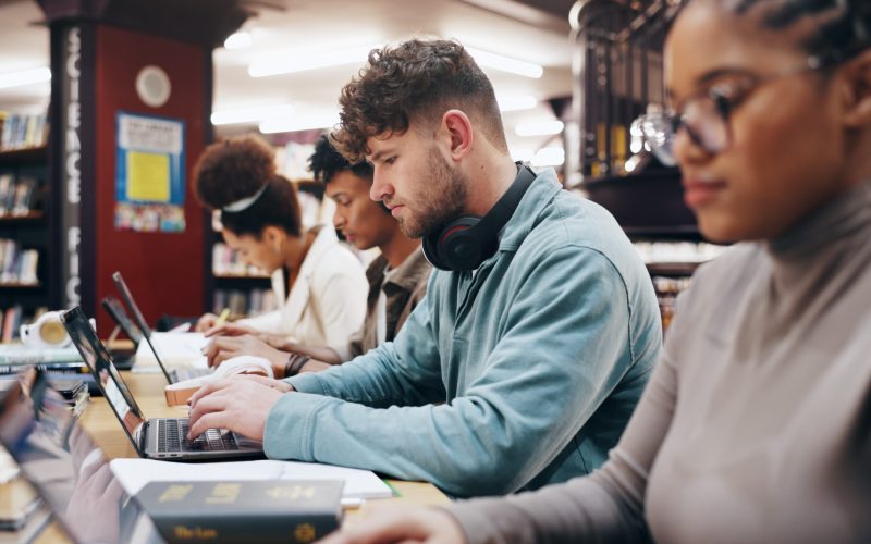 Education, laptop and typing with student friends in library of university together for learning or study. Computer, development or growth with serious men and women on college campus for scholarship.